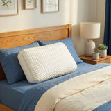 The Pillowash Crisp pillow propped against blue linen bedding on an oak bed, beside a lamp and a potted plant