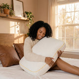 A woman sits on her bed in morning light hugging the Pillowash Crisp pillow to her chest