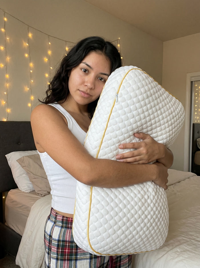 Young woman in a white tank top hugging a quilted Pillowash washable pillow in a bedroom lit by string lights